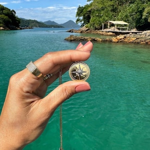 Könnte beinhalten: Eine goldfarbene Kompass-Anhänger-Halskette mit Stern-Design, gehalten vor türkisfarbenem Wasser. Der Anhänger ist rund mit einer detaillierten Windrose. Ein goldener Ring mit kleinen Steinen befindet sich am Finger der Person.