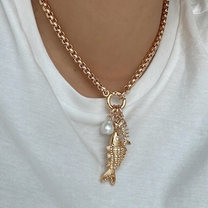May include: Gold-coloured necklace with a chunky chain and a pendant. The pendant includes a fish charm, a pearl, and other small charms. The fish charm has detailed features. The necklace is worn against a white background.