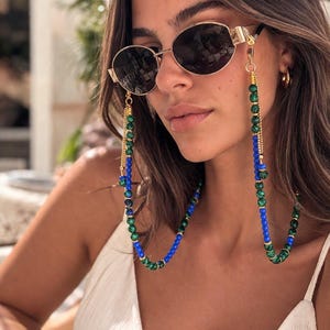 Puede incluir: Gafas de sol redondas con montura dorada y una cadena de cuentas. La cadena presenta cuentas verdes y azules alternas, con detalles dorados. Las gafas de sol tienen lentes oscuras.