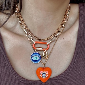 Puede incluir: Un collar dorado en capas con un mosquetón naranja, un colgante de ojo azul y blanco, un dije de concha dorada y un colgante de corazón naranja con un diseño de ojo. El collar se lleva puesto.