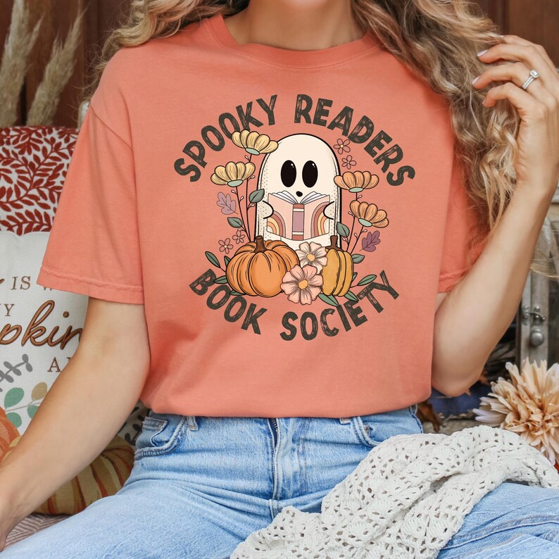 Pode incluir: Uma camiseta cor de p&ecirc;ssego com um gr&aacute;fico de um fantasma lendo um livro cercado por flores e ab&oacute;boras. O texto "Spooky Readers Book Society" est&aacute; impresso na camiseta.