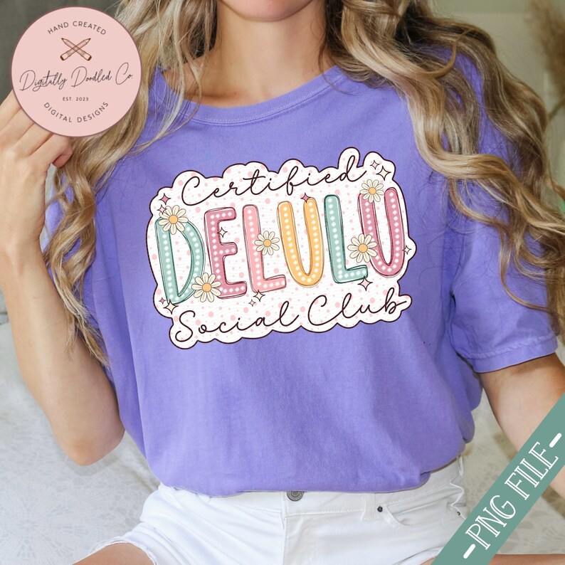 May include: A purple t-shirt with a white graphic design that says "Certified Delulu Social Club". The graphic design features a colorful, outlined word "Delulu" with white polka dots and flowers.