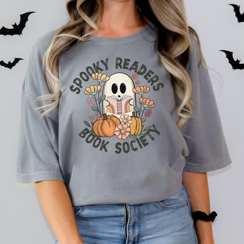 Pode incluir: Uma camiseta cinza com um gr&aacute;fico de um fantasma lendo um livro cercado por flores e ab&oacute;boras. O texto "Spooky Readers Book Society" est&aacute; impresso na camiseta.