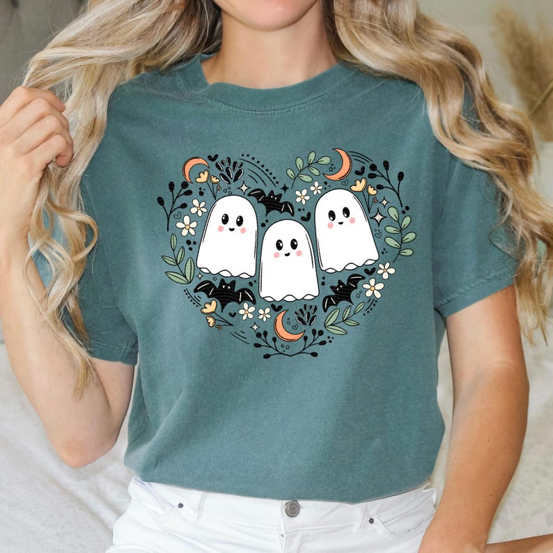Op de afbeelding: Tealblauw T-shirt met een wit hartvormig ontwerp met drie cartoon-geesten met glimlachende gezichten en vleermuizen. Het ontwerp is omringd door bloemen en bladeren.