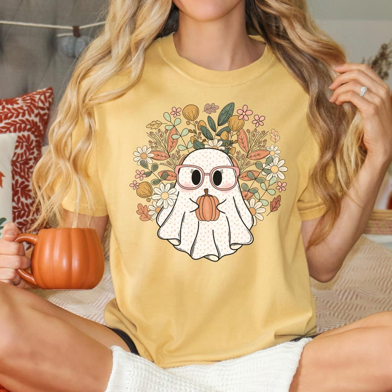 May include: A yellow t-shirt with a white ghost wearing pink glasses and holding a small orange pumpkin. The ghost is surrounded by a floral design with pink, yellow, and green flowers.