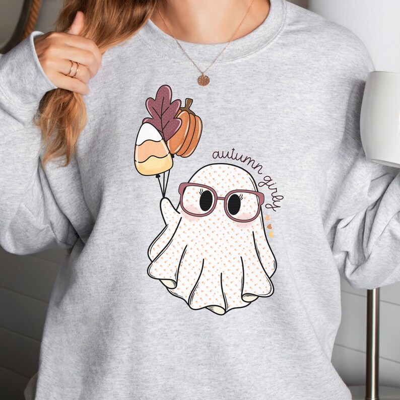 May include: A grey sweatshirt featuring a white ghost wearing glasses and holding a candy corn and a pumpkin. The text "autumn girly" is written on the sweatshirt.