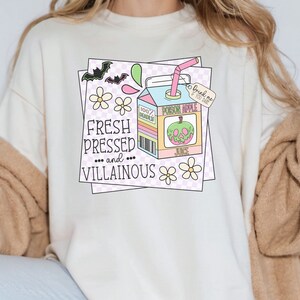 May include: A white t-shirt with a graphic of a cartoon juice box with a green apple with a skull face. The text on the juice box says "Poison Apple Juice". The text on the shirt says "Fresh Pressed... and... Villainous".