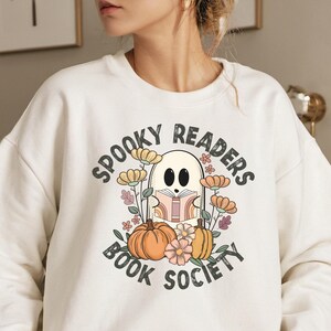 Pode incluir: Camisola branca com um gr&aacute;fico de um fantasma a ler um livro rodeado de flores e ab&oacute;boras. O texto "Spooky Readers Book Society" est&aacute; escrito em torno do fantasma.