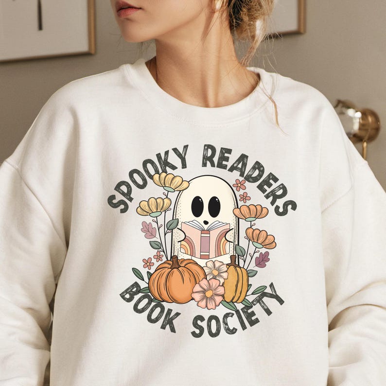 Pode incluir: Camisola branca com um gr&aacute;fico de um fantasma a ler um livro rodeado de flores e ab&oacute;boras. O texto "Spooky Readers Book Society" est&aacute; escrito em torno do fantasma.
