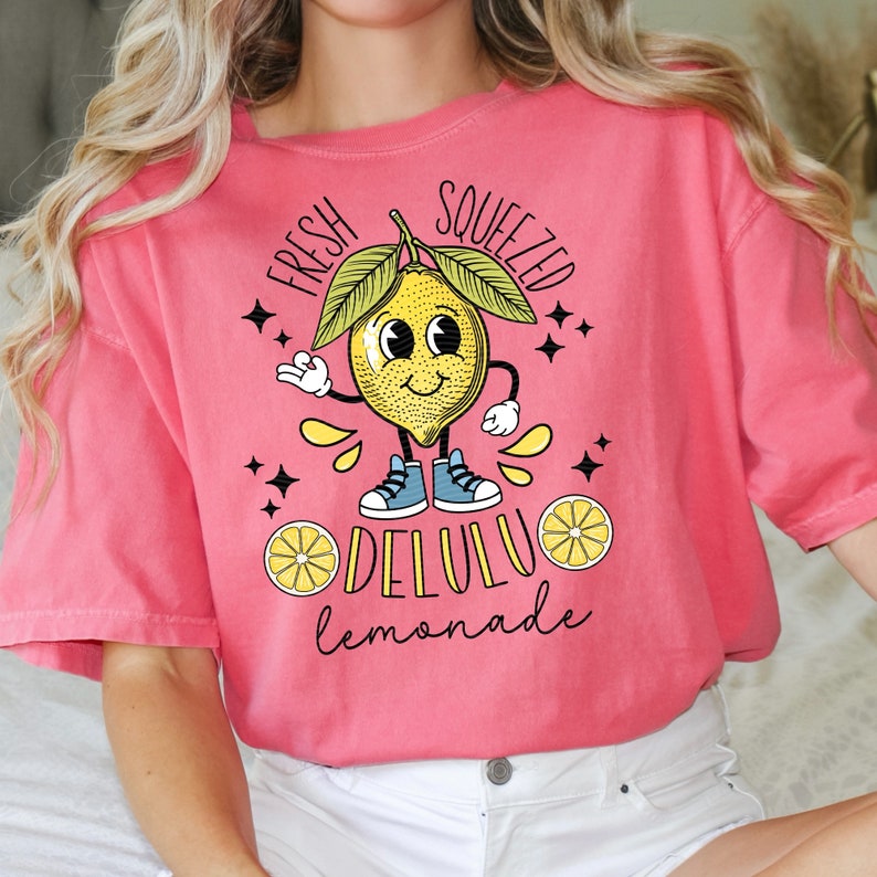 Pu&ograve; includere: Una maglietta rosa con un limone a cartone animato e il testo "Fresh Squeezed Delulu Lemonade".