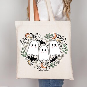Op de afbeelding: Een witte canvas tote bag met een zwart-witte illustratie van drie geesten in een hartvorm omringd door vleermuizen, bloemen en manen. De geesten hebben zwarte ogen en glimlachen.