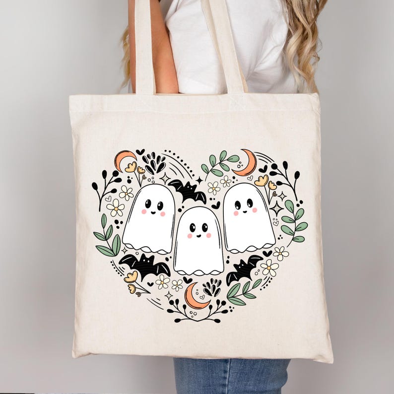 Op de afbeelding: Een witte canvas tote bag met een zwart-witte illustratie van drie geesten in een hartvorm omringd door vleermuizen, bloemen en manen. De geesten hebben zwarte ogen en glimlachen.