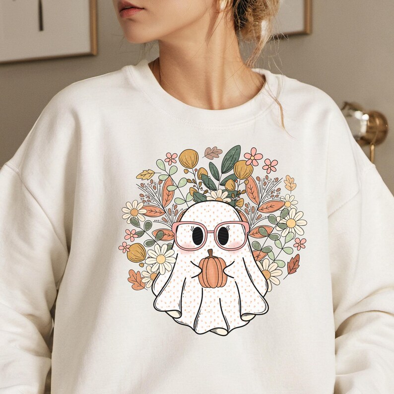 May include: A white sweatshirt with a cute cartoon ghost wearing glasses and holding a pumpkin. The ghost is surrounded by a wreath of fall leaves and flowers.