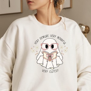 May include: White sweatshirt featuring a cute cartoon ghost wearing pink glasses and reading a book. The ghost is surrounded by small pink hearts and white flowers. The text on the sweatshirt reads "Very Demure Very Mindful Very Cutesy".