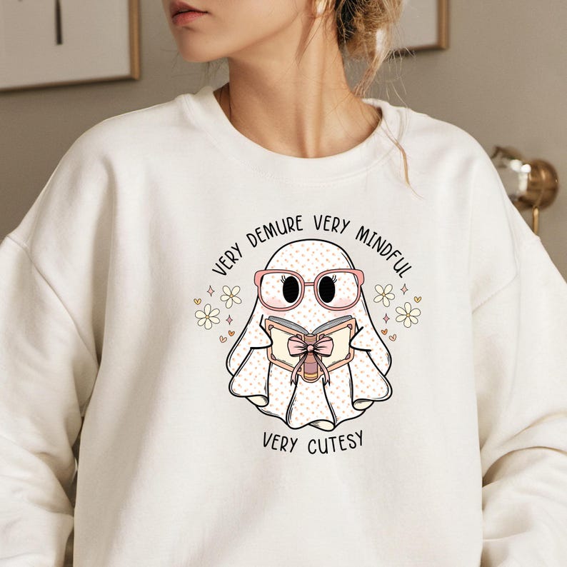 May include: White sweatshirt featuring a cute cartoon ghost wearing pink glasses and reading a book. The ghost is surrounded by small pink hearts and white flowers. The text on the sweatshirt reads "Very Demure Very Mindful Very Cutesy".