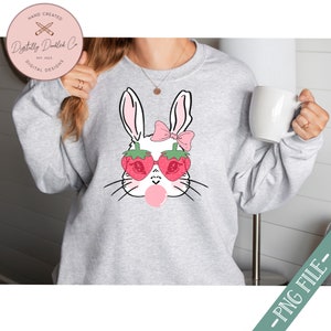 May include: A white bunny with pink ears and a pink bow wearing strawberry-shaped sunglasses and blowing a pink bubble. The bunny is on a gray sweatshirt.