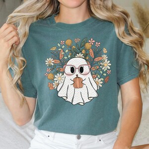 May include: Teal blue t-shirt with a white ghost wearing pink glasses and holding a small orange pumpkin. The ghost is surrounded by a floral design with pink, yellow, and green flowers.