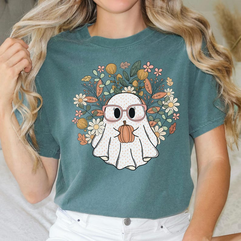May include: Teal blue t-shirt with a white ghost wearing pink glasses and holding a small orange pumpkin. The ghost is surrounded by a floral design with pink, yellow, and green flowers.