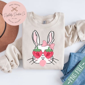 May include: A white bunny with pink bow and strawberry sunglasses blowing a bubble. The bunny is on a beige sweatshirt.