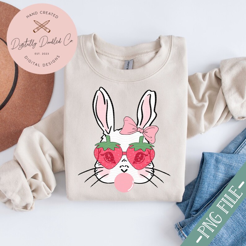 May include: A white bunny with pink bow and strawberry sunglasses blowing a bubble. The bunny is on a beige sweatshirt.