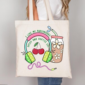 May include: A white tote bag with a colorful graphic design featuring headphones, cherries, and a glass of iced coffee. The text on the bag reads "I like my audiobooks spicy and coffee icy."