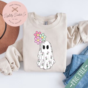 May include: A white ghost with a floral pattern holding a bouquet of pink, blue, and green flowers. The ghost is on a beige sweatshirt.