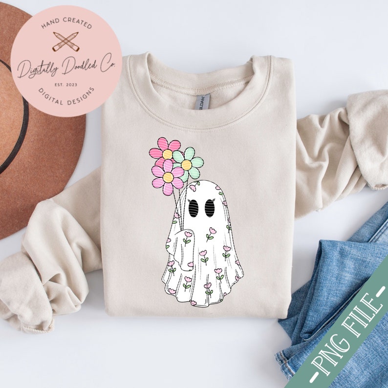 May include: A white ghost with a floral pattern holding a bouquet of pink, blue, and green flowers. The ghost is on a beige sweatshirt.