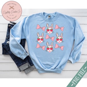 May include: Light blue sweatshirt with a repeating pattern of white bunnies wearing pink bows and strawberry sunglasses. The bunnies are holding pink lollipops.