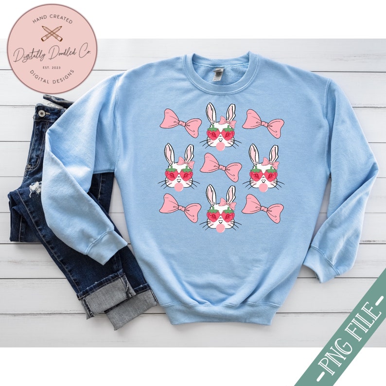 May include: Light blue sweatshirt with a repeating pattern of white bunnies wearing pink bows and strawberry sunglasses. The bunnies are holding pink lollipops.