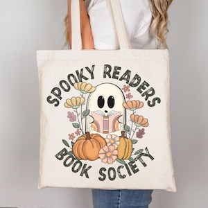 Pode incluir: Uma bolsa de tecido branca com o texto "Spooky Readers Book Society" em preto. Um fantasma de desenho animado est&aacute; segurando um livro com p&aacute;ginas arco-&iacute;ris e est&aacute; rodeado de flores e ab&oacute;boras.