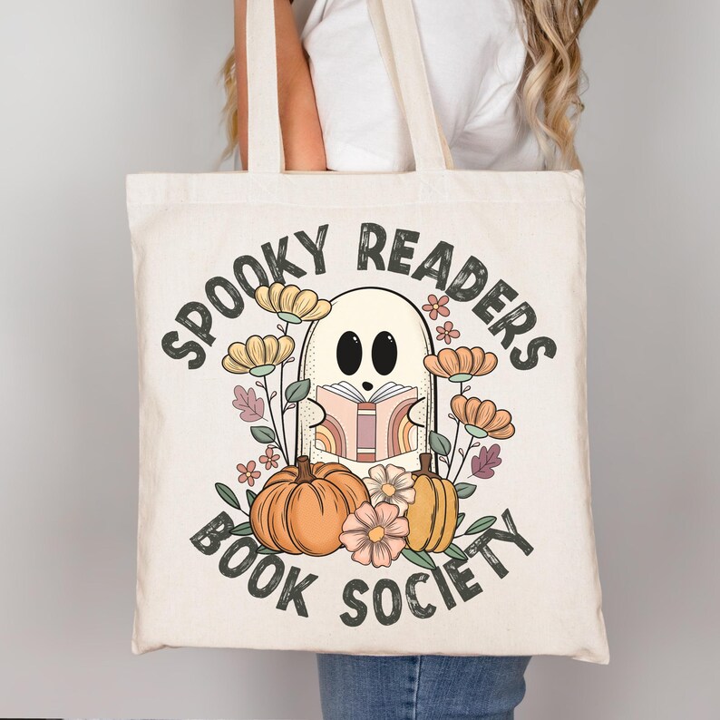 Pode incluir: Uma bolsa de tecido branca com o texto "Spooky Readers Book Society" em preto. Um fantasma de desenho animado est&aacute; segurando um livro com p&aacute;ginas arco-&iacute;ris e est&aacute; rodeado de flores e ab&oacute;boras.