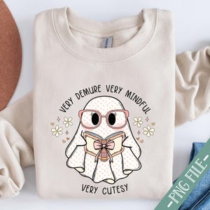 May include: A beige sweatshirt featuring a cute ghost illustration wearing glasses and reading a book. The text on the sweatshirt reads "Very Demure Very Mindful Very Cutesy".