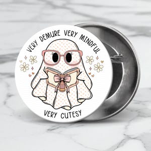 May include: A white ghost with pink glasses and a pink bow on a book is reading. The ghost is surrounded by white flowers and hearts. The text around the ghost says "Very Demure Very Mindful Very Cutesy".
