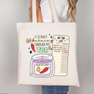 May include: A white tote bag with a graphic of a candle and a list titled "My TBR List". The candle has a label that says "Caution Spicy Content". The list has a column for "Title" and "Spice Level". The graphic is in a cartoon style with bright colors.