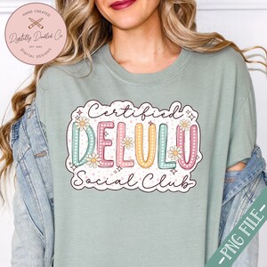May include: A light green t-shirt with a graphic design that says "Certified Delulu Social Club". The graphic is in a colorful, retro style with a daisy pattern and white polka dots.
