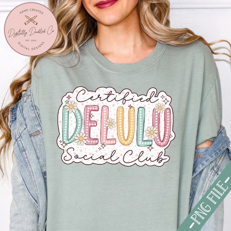 May include: A light green t-shirt with a graphic design that says "Certified Delulu Social Club". The graphic is in a colorful, retro style with a daisy pattern and white polka dots.