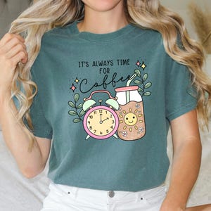 May include: Teal blue t-shirt with a graphic design of a coffee cup with a smiling sun face, a straw, and an alarm clock. The text on the shirt reads "It's always time for coffee."