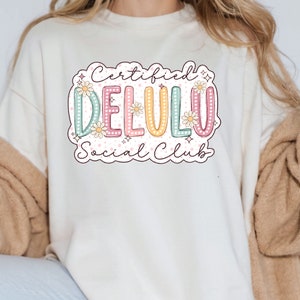 May include: A white sweatshirt with a graphic design that says "Certified Delulu Social Club" in a colorful, retro-inspired font with flowers and stars.