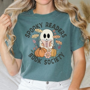 Pode incluir: Camiseta azul turquesa com um gr&aacute;fico de um fantasma lendo um livro cercado por flores e ab&oacute;boras. O texto "Spooky Readers Book Society" est&aacute; impresso na camiseta.