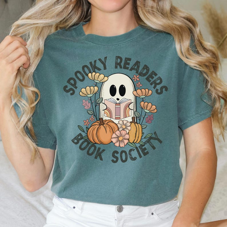 Pode incluir: Camiseta azul turquesa com um gr&aacute;fico de um fantasma lendo um livro cercado por flores e ab&oacute;boras. O texto "Spooky Readers Book Society" est&aacute; impresso na camiseta.