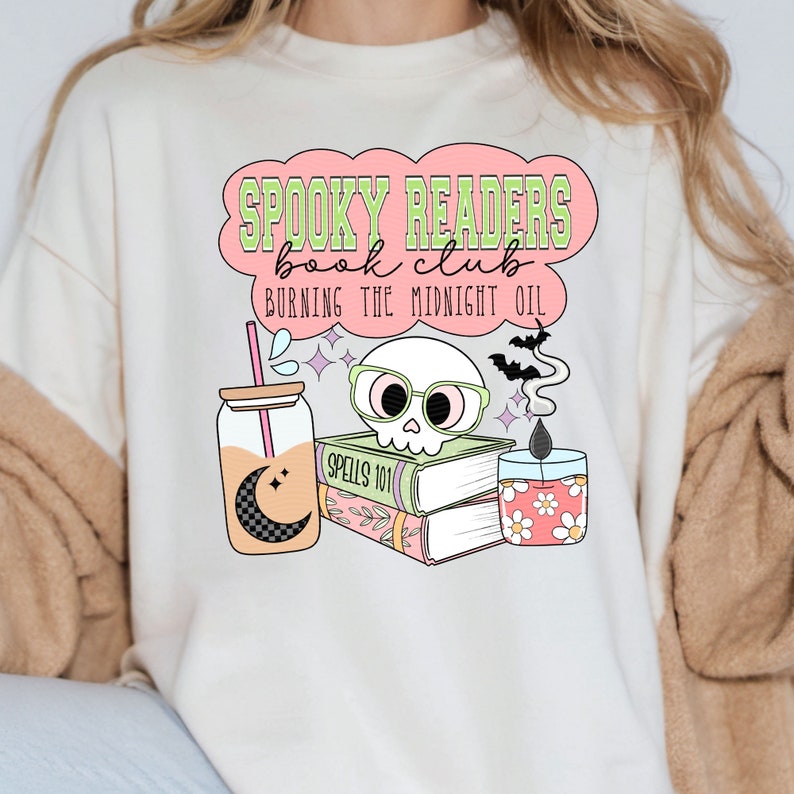 May include: A white t-shirt with a pink and green graphic design that says "Spooky Readers Book Club Burning the Midnight Oil". The graphic includes a skull wearing glasses, a stack of books, a candle, and a glass jar with a straw.