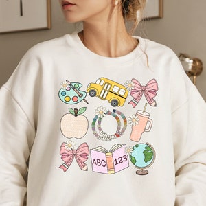 May include: Cream-colored sweatshirt featuring a colorful school-themed graphic. The design includes a yellow school bus, a pink bow, an apple, a palette, a book with "ABC 123", a globe, and a tumbler. The word "TEACH" is on a bracelet.