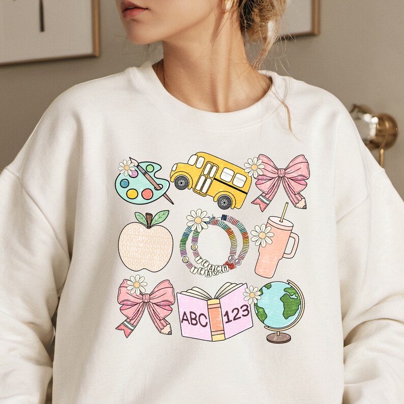 May include: Cream-colored sweatshirt featuring a colorful school-themed graphic. The design includes a yellow school bus, a pink bow, an apple, a palette, a book with "ABC 123", a globe, and a tumbler. The word "TEACH" is on a bracelet.