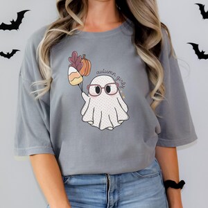 May include: A grey t-shirt featuring a cute ghost wearing glasses and holding a bunch of autumnal items. The text "autumn girly" is written on the shirt.