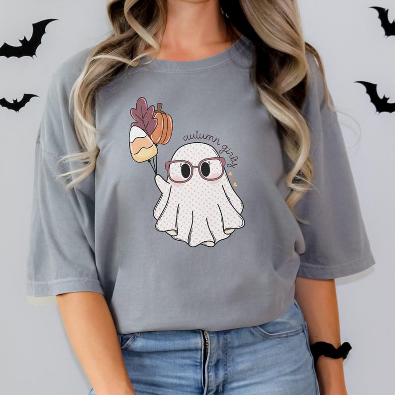 May include: A grey t-shirt featuring a cute ghost wearing glasses and holding a bunch of autumnal items. The text "autumn girly" is written on the shirt.