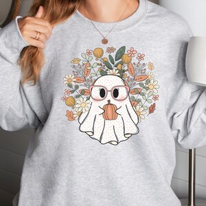 May include: A gray sweatshirt with a white ghost wearing pink glasses and holding a small orange pumpkin. The ghost is surrounded by a wreath of colorful flowers and leaves.