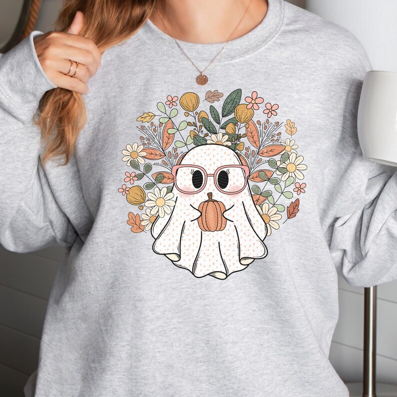 May include: A gray sweatshirt with a white ghost wearing pink glasses and holding a small orange pumpkin. The ghost is surrounded by a wreath of colorful flowers and leaves.