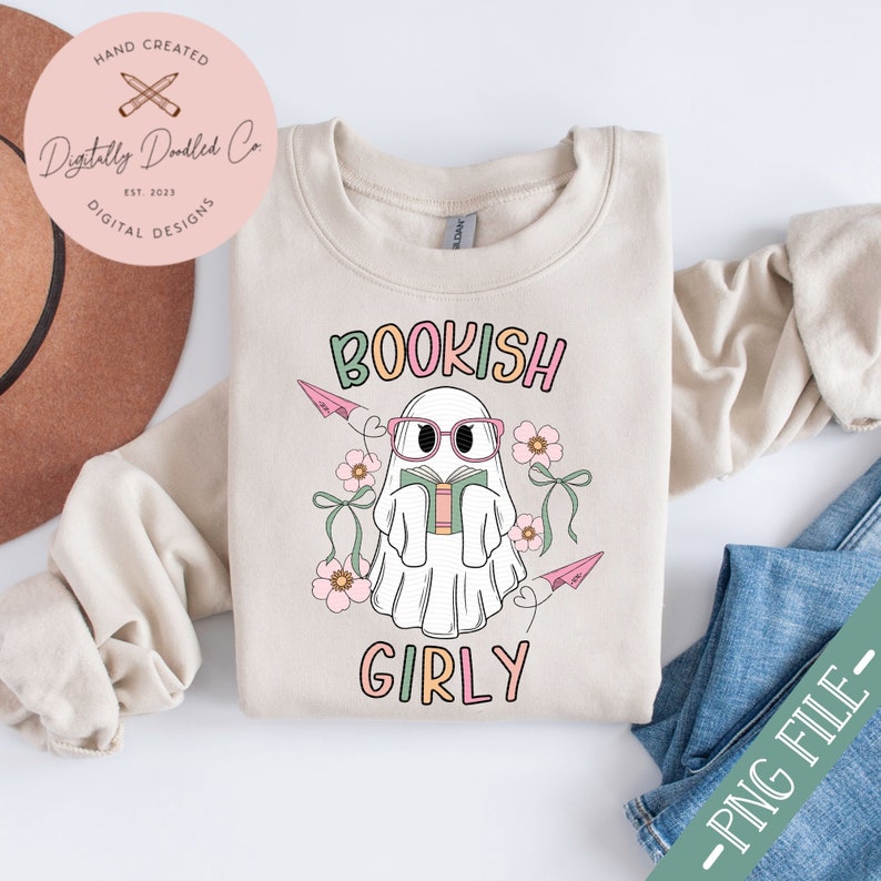 K&ouml;nnte beinhalten: Ein beiger Pullover mit einem wei&szlig;en Geist, der eine rosa Brille tr&auml;gt und ein Buch liest. Der Geist ist von rosa Blumen und Papierflugzeugen umgeben. Der Text "BOOKISH GIRLY" ist auf dem Pullover aufgedruckt.