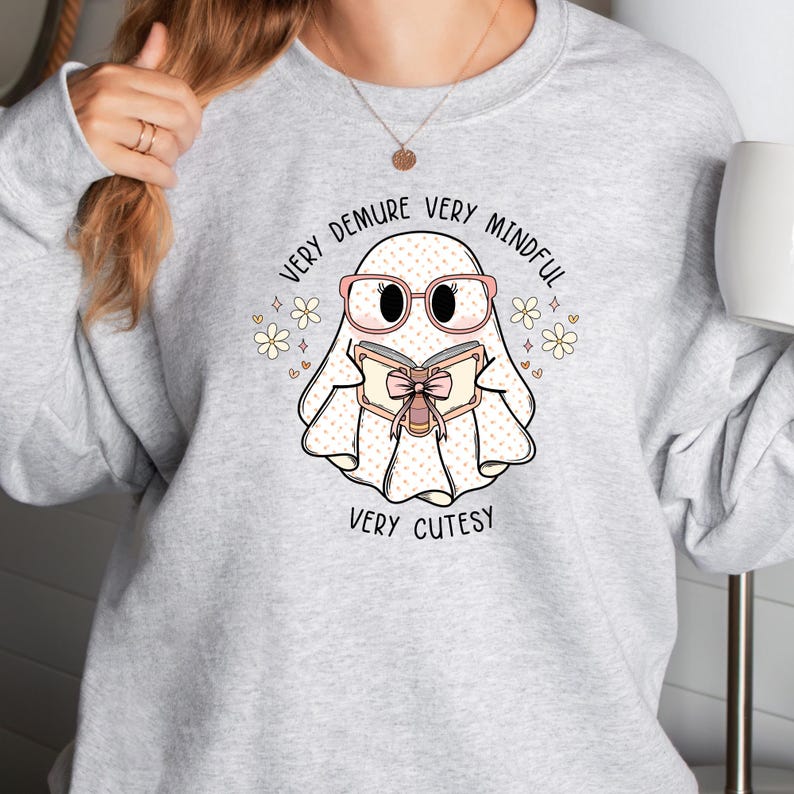 May include: A grey sweatshirt featuring a cute ghost wearing pink glasses and reading a book. The ghost has pink polka dots and the text reads "Very Demure Very Mindful Very Cutesy".