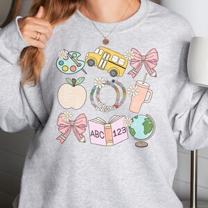 May include: A light gray sweatshirt featuring colorful school-themed illustrations. The design includes a yellow school bus, a pink bow with a pencil, an apple, a book with "ABC 123", a globe, and a paint palette. The sweatshirt has a crew neck.